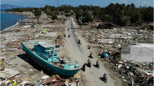 People drive past a washed up boat and collapsed buildings in Palu on October 1, 2018, after an earthquake and tsunami hit the area on September 28