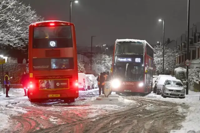 Два красных лондонских автобуса на небольшом перекрестке, дорога в снегу, горят фонари, дома по обеим сторонам
