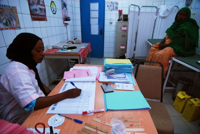 Une femme assise dans un bureau en train d'écrire, elle porte une blouse violette, une autre femme est assise sur un lit en face d'elle.