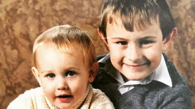Una foto antigua de dos chicos. El de la izquierda tiene el pelo rubio y un jersey de lana color crema. El de la derecha es mayor, con el pelo corto y claro, camisa blanca y jersey gris. Están sentados frente a un fondo marrón.