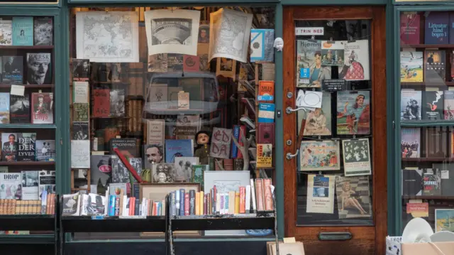 通り沿いの書店のウィンドウや入口にポスターが貼られ、ディスプレイにさまざまな本が並べられている。ウィンドウ越しに店内の書棚も見える。店の外にも低い棚が置かれ、本が並べられている