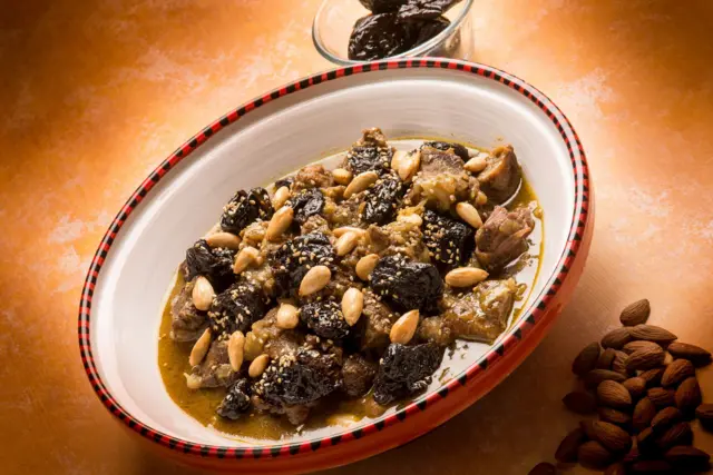 حصة من طاجين اللحم والبرقوق وضعت بطبق مزخرف الحواف أعلى منضدة وقد اكتسبت لونا بنيا. داخل الطبق مكعبات لحم مطهوة ورشّت باللوز والسمسم وسط سائل بسيط