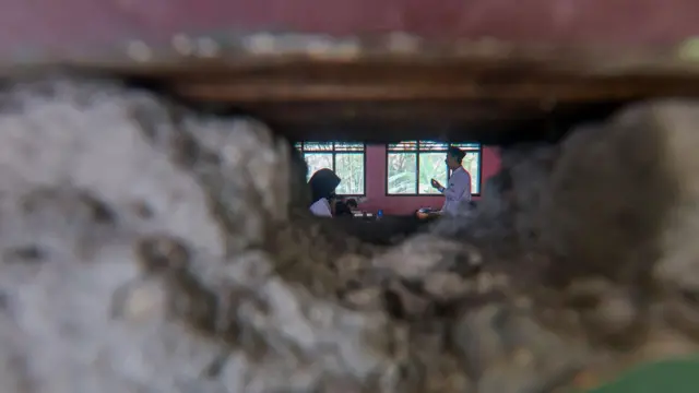 Seorang guru tampak sedang mengajar diintip kamera dari tembok yang jebol.