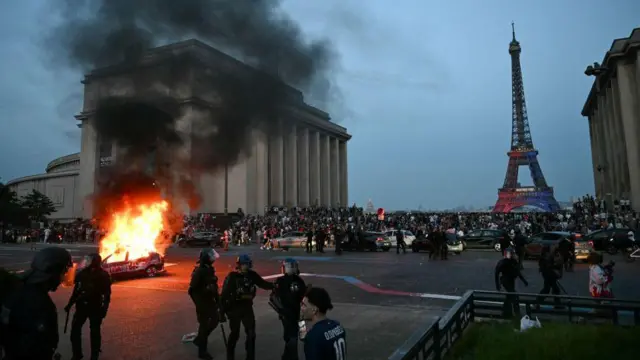 A la izquierda se ve un vehículo en llamas. Muy cerca está un grupo de policías. Al fondo se ven vehículos de la policía, una concentración de personas y la Torre Eiffel