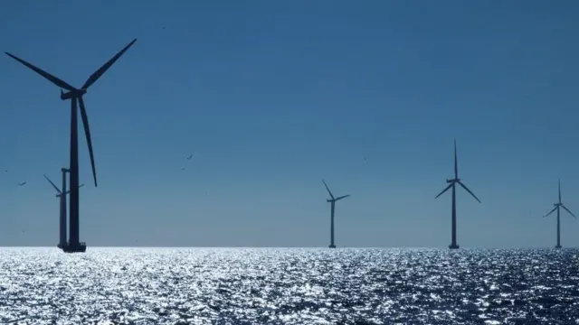 Cinco turbinas eólicas en silueta salpicadas sobre un mar en calma, contra un cielo azul.