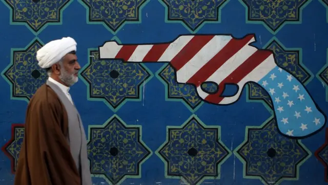 An Iranian clerric walks past a mural on the wall of the former US embassy