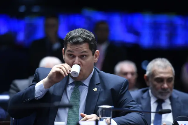 Davi Alcolumbre toma caféda para jogar na lotofacil onlineuma pequena xícara branca no plenário do Senado. Ele está vestido com um terno azul marinho, camisa azul clara e gravata turquesa