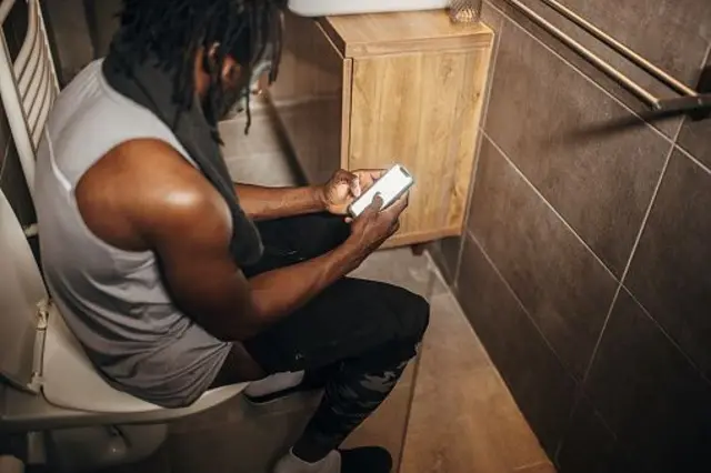 Un homme noir assis sur les toilettes et utilisant son smartphone dans la salle de bain le matin.