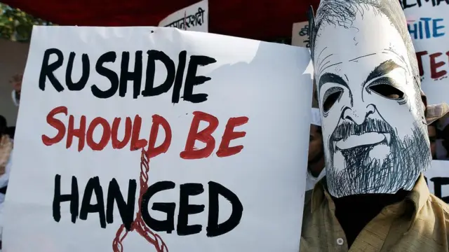 Un musulman indien porte un masque de l'écrivain indien Salman Rushdie alors qu'il affiche une pancarte condamnant Rushdie lors d'une manifestation à Bombay, le 12 janvier 2004. Lors de la manifestation contre la présence de Rushdie dans la ville, organisée par plusieurs organisations musulmanes, une récompense de 100 000 roupies (2 199 USD) a été offerte à quiconque noircira le visage de Rushdie, qui, dans son livre Les Versets sataniques, aurait tenu des propos contre le saint prophète Mahomet.