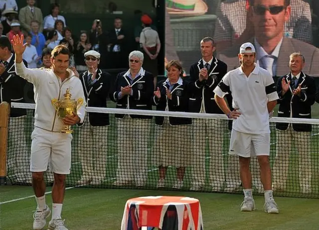 Federer celebrates his 2009 Wimbledon title, while a TV screen shows Sampras watching on from the stands