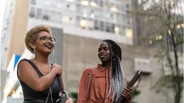 Fotografia colorida mostra duas mulheres jovens e negras; uma tem cabelo comprido e segura um caderno, a outra tem cabelo curto e usa óculos, ambas sorriem