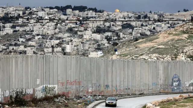 Carro passa por muro israelense na cidade palestinapixbet portugalAbu Dis, com Jerusalém ao fundo