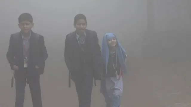 Dos niños y una niña caminando hacia la escuela en medio de una neblina gris de contaminación.