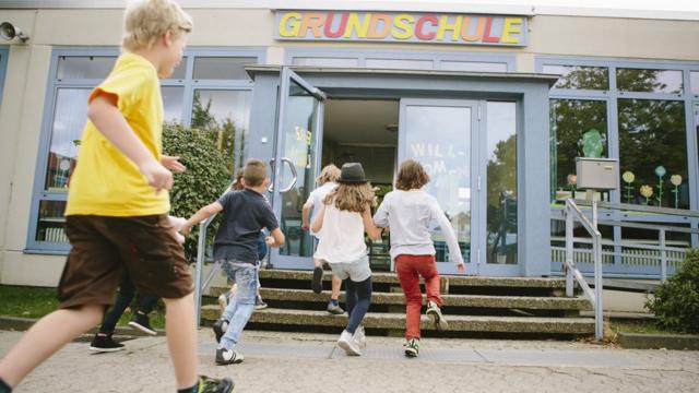 Crianças correm para entrar em um edifício, onde está escrito 'Grundschule' (escola primária, em alemão)