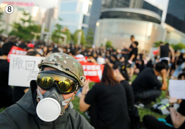 A man in a yellow sun visor and wearing what looks like a gas mask looks at teh camera, while behind him a large crowd of