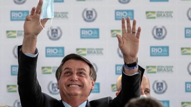 O presidente Jair Bolsonaro sorri e levanta as mãos para o alto; em uma delas segura uma máscara