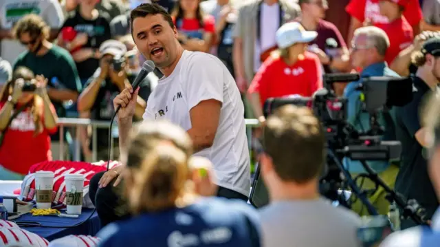 Charlie Kirk, vestido con una camiseta blanca, hablando a las cámaras en el evento donde le dispararon.
