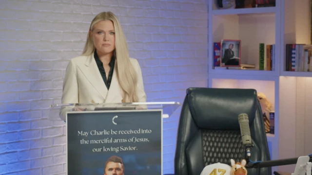 Erika Kirk kocasının podcast yaptığı koltuğun yanında bir kürsüde konuşuyor.