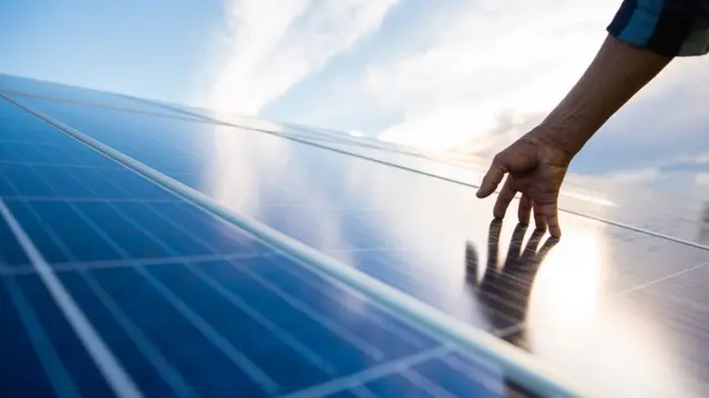 Mão toca um painel fotovoltaico contra um céu azul com nuvens