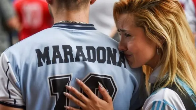 A woman rests on the shoulder of a man wearing a Maradona football shirt