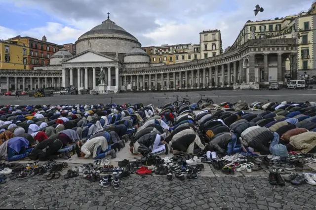 Десятки верующих стоят на коленях, склонив головы к земле в молитве, десятки пар обуви разбросаны на обочине у площади Плебисцито