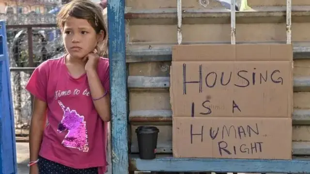 ਗਰੀਸ ਵਿੱਚ ਘਰ ਇੱਕ ਮਨੁੱਖੀ ਹੱਕ ਹੈ ਦੇ ਪੋਸਟਰ ਕੋਲ ਖੜ੍ਹੀ ਇੱਕ ਸ਼ਰਣਾਰਥੀ ਕੁੜੀ