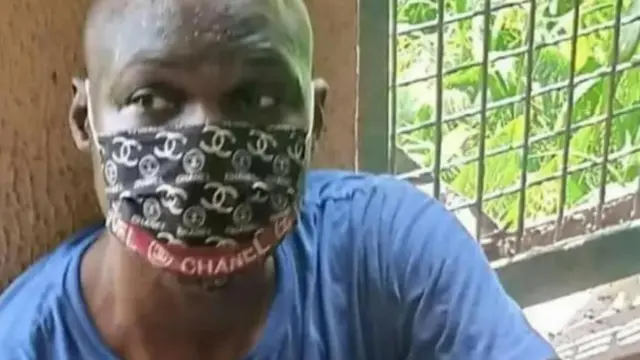 Baba Ijesha nínú àhámọ́ ọgbà ẹ̀wọ̀n, ó joko silẹ, ó fi í ọmú bo ẹnu rẹ, o si wọ aṣọ aláwọ̀ búlúù. 