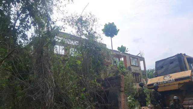 Nnukwu ụgbọ katapila nọ n'akụkụ ọhịa na ndị ọrụ nchekwa nọ ha n'akụkụ.