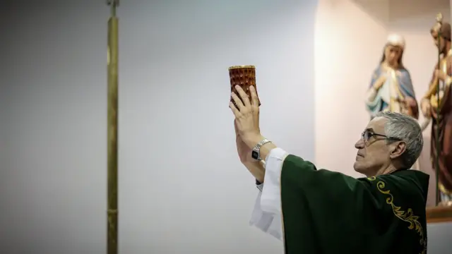 Liciorola roletaperfil erguendo objeto sagrado na missa; ao fundo, imagensrola roletasantos