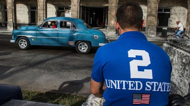 Un hombre de espaldas con una camiseta de Estados Unidos (en inglés) y la bandera de ese país en Cuba.