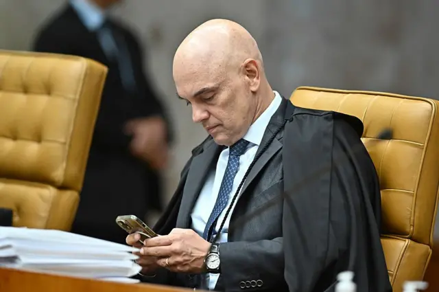 Moraes sentado em plenário do STF, olhando para celular