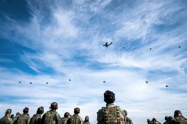 Кільканадцять військових спостерігають за десантуванням з літака: у небі видно літак і близько десяти рівномірно розподілених на відстані парашутів.