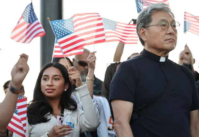 Varias personas ondean banderas estadounidenses, entre ellos un cura vestido con sotana en primer plano y una joven con pelo negro detrás, durante una vigilia en honor a Narciso Barranco y en protesta por las redadas migratorias del gobierno de Donald Trump, en Santa Ana, California, Estados Unidos, el 27 de junio de 2025. (Foto: Mario Tama/Getty Images)
