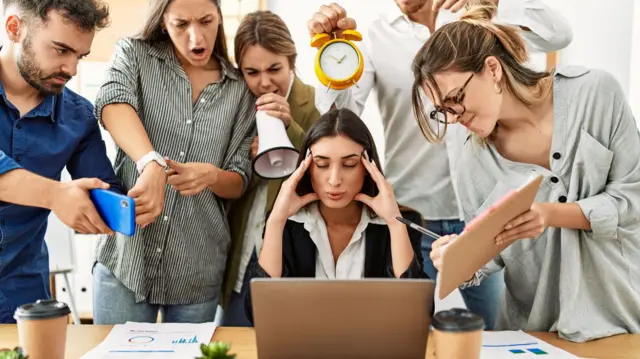 مجموعة من الموظفين يصرخون في وجه زميلة لهم تجلس أمام اللابتوب وقد أغمضت عينيها ووضعت يديها على وجنتيها وتبدو وكأنها تحاول التنفس بعمق.