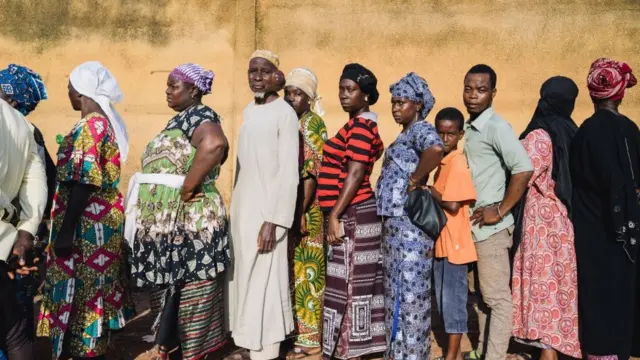 Mata da tsirarun maza a kan layi. Sanye da tufafi launi daban-daban. Wasu daga ciki na kallon kamara, wasu su na kallon gabansu wasu sun bata rai.