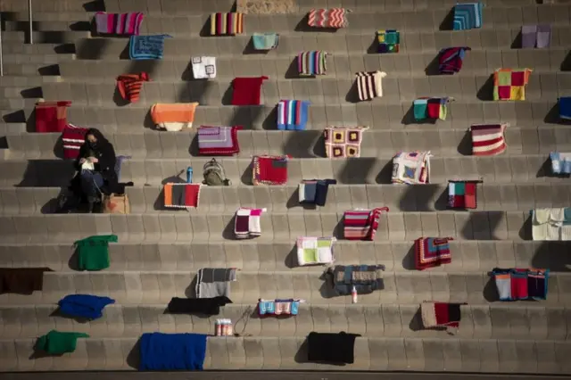 A women knits a blanket while she sits among hundreds of other blankets.