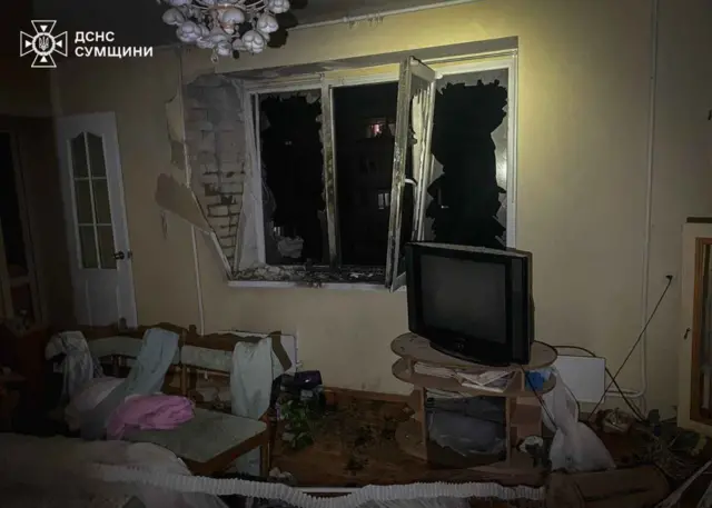 Кімната в приватному будинку після вибуху: вікно вибите, скло розсипане всередині, пошкоджена стіна. У приміщенні стоять диван і стільці, поруч телевізор, на підлозі уламки й речі, видні двори багатоповерхівок за розбитим вікном.