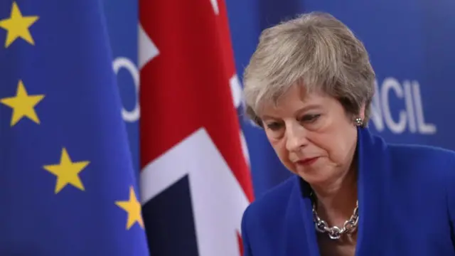 Theresa Mayesportenet com betfrente a bandeiras do Reino Unido e da União Europeia