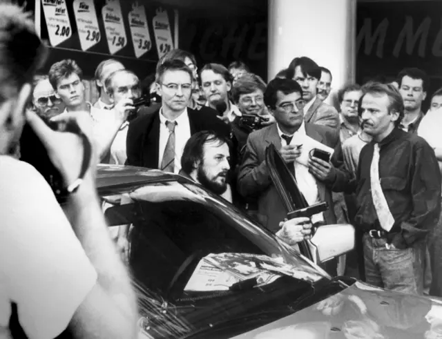 Koyu bir ceket giyen ve kravat takan Udo Röbel, elinde silah tutan Hans Juergen Rösner'in hemen arkasında duruyor.