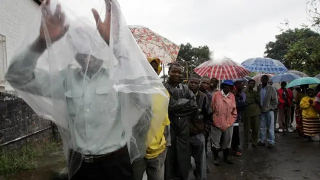 People line up inside rain with umbrella. One man use plastic bag cover imsef