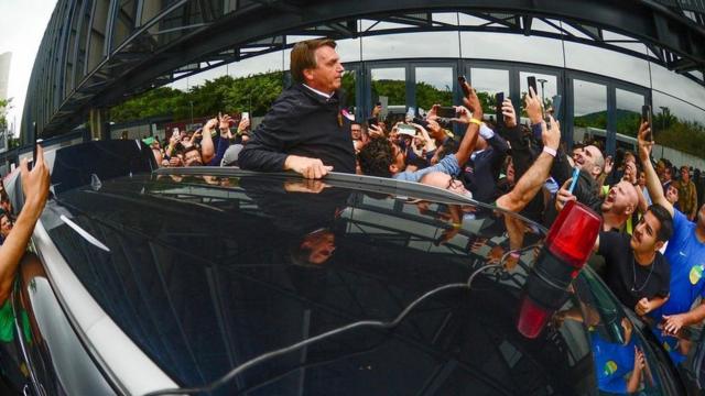 Bolsonaro em pé para fora do carro, enquanto pessoas tentam tirar foto com ele