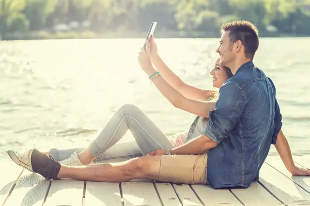 Pareja sentada en un muelle tomándose una foto con un celular.