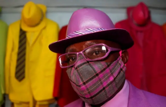 Kenyan fashionista James Maina Mwangi, dressed all in purple, stands beside green, yellow and red suits.