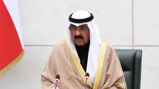 أمير الكويت، الشيخ مشعل الأحمد الجابر الصباح واقفا وهو يرتدي ثوبا أسودا وعليه مشلح بني فاتح اللون ويضع على رأسه شماغ أبيض وعقال أسود.
