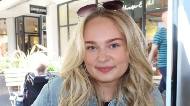 Kobieta o blond włosach patrzy w kamerę i uśmiecha się. Ma na sobie dżinsową kurtkę i okulary przeciwsłoneczne. Tło jest lekko rozmyte, widać w nim inne osoby i witryny sklepów.