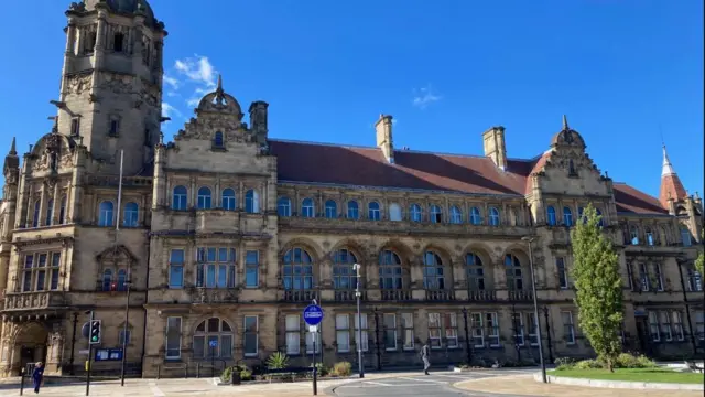 Ayuntamiento del Condado de Wakefield. Un gran edificio ornamentado de tres plantas con ventanas arqueadas, techo de tejas rojas y torretas a ambos lados. El cielo azul se alza sobre él.