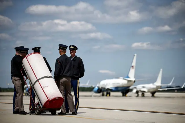 تیمی از پرسنل امنیتی روی باند فرودگاه پایگاه مشترک اندروز، مریلند، ایستاده‌اند و فرش سرخ پیچیده ‌شده‌ای را جلوی هواپیماهای آمریکایی نگه داشته‌اند و منتظر ورود هواپیمای سلطنتی در روز دوشنبه هستند.