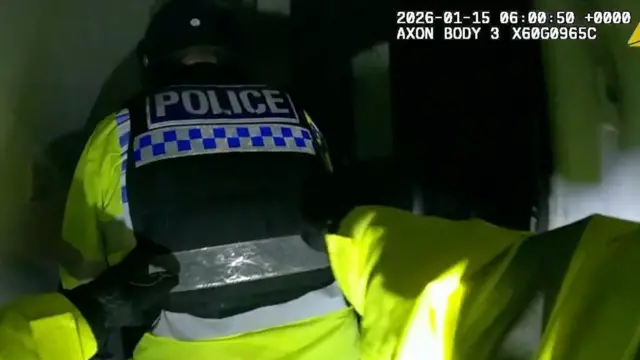 En la esquina superior derecha se encuentra la fecha y hora de la cámara corporal de la policía. La imagen muestra la espalda de un agente de policía con una chaqueta de alta visibilidad y un chaleco negro encima, con la palabra POLICÍA escrita en blanco en la espalda. Se ven los brazos de dos de sus compañeros en primer plano. El fondo está desenfocado, pero es el recibidor de una casa.