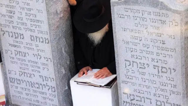 Rabino agachado diante de livro sagrado, entre dois túmulos