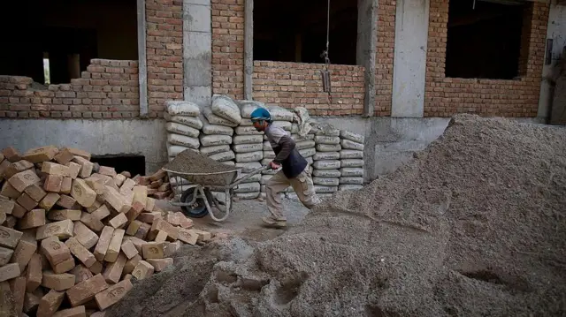 یک ساختمان نیمه‌کاره که یک کارگر در حال انتقال در یک کراچی است و انبار ریگ، خشت و خریطه‌های سمنت دیده می‌شود.